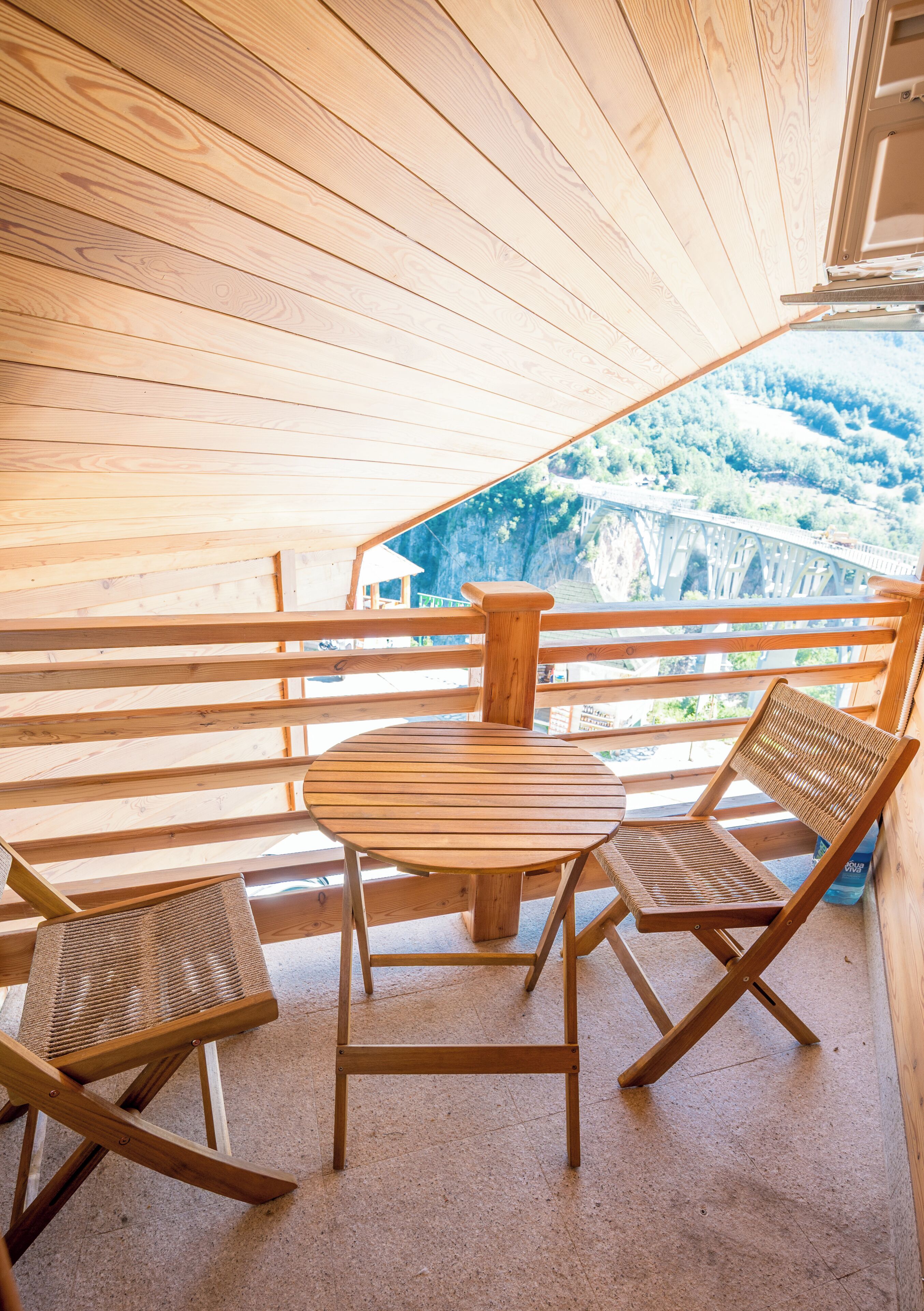 Una terraza de madera con una mesa redonda y dos sillas, con vista a un paisaje que incluye un puente y un bosque.
