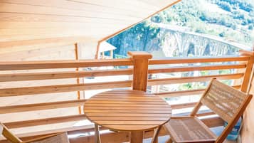Una terraza de madera con una mesa redonda y dos sillas, con vista a un paisaje que incluye un puente y un bosque.