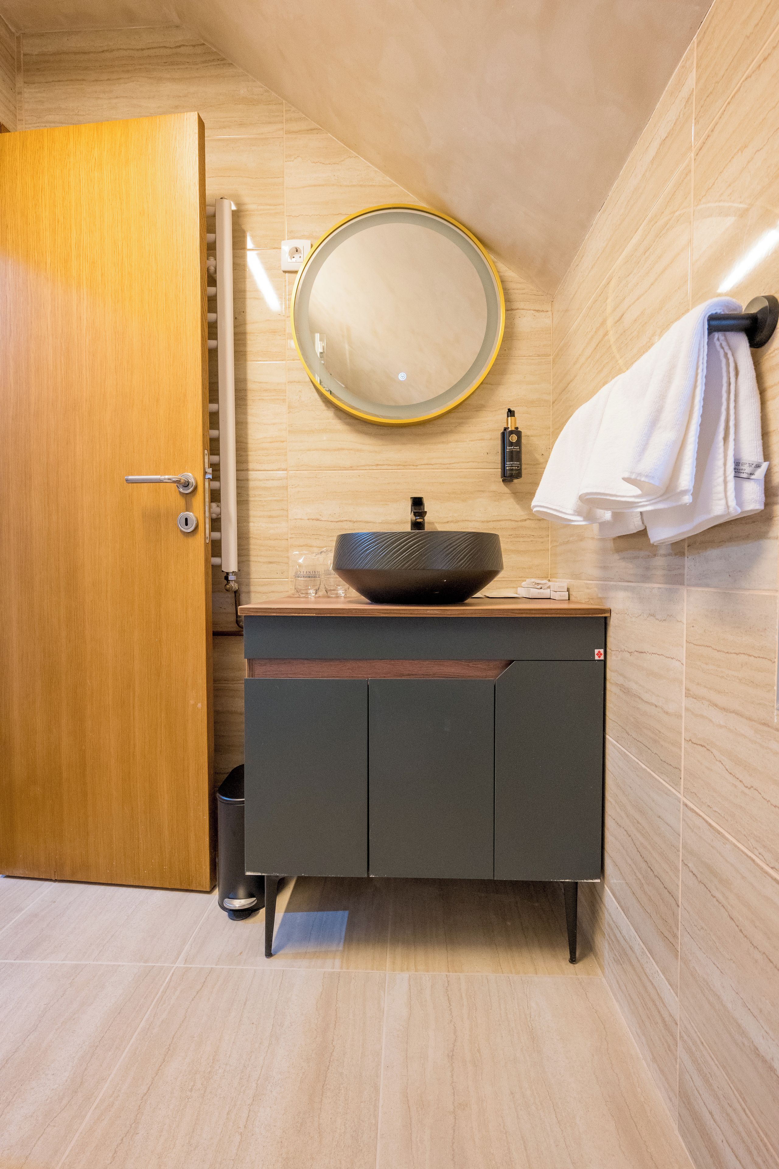 Een badkamer met een houten deur, een ronde spiegel, een wastafel met een bijzondere waskom en een handdoekrek met een witte handdoek.