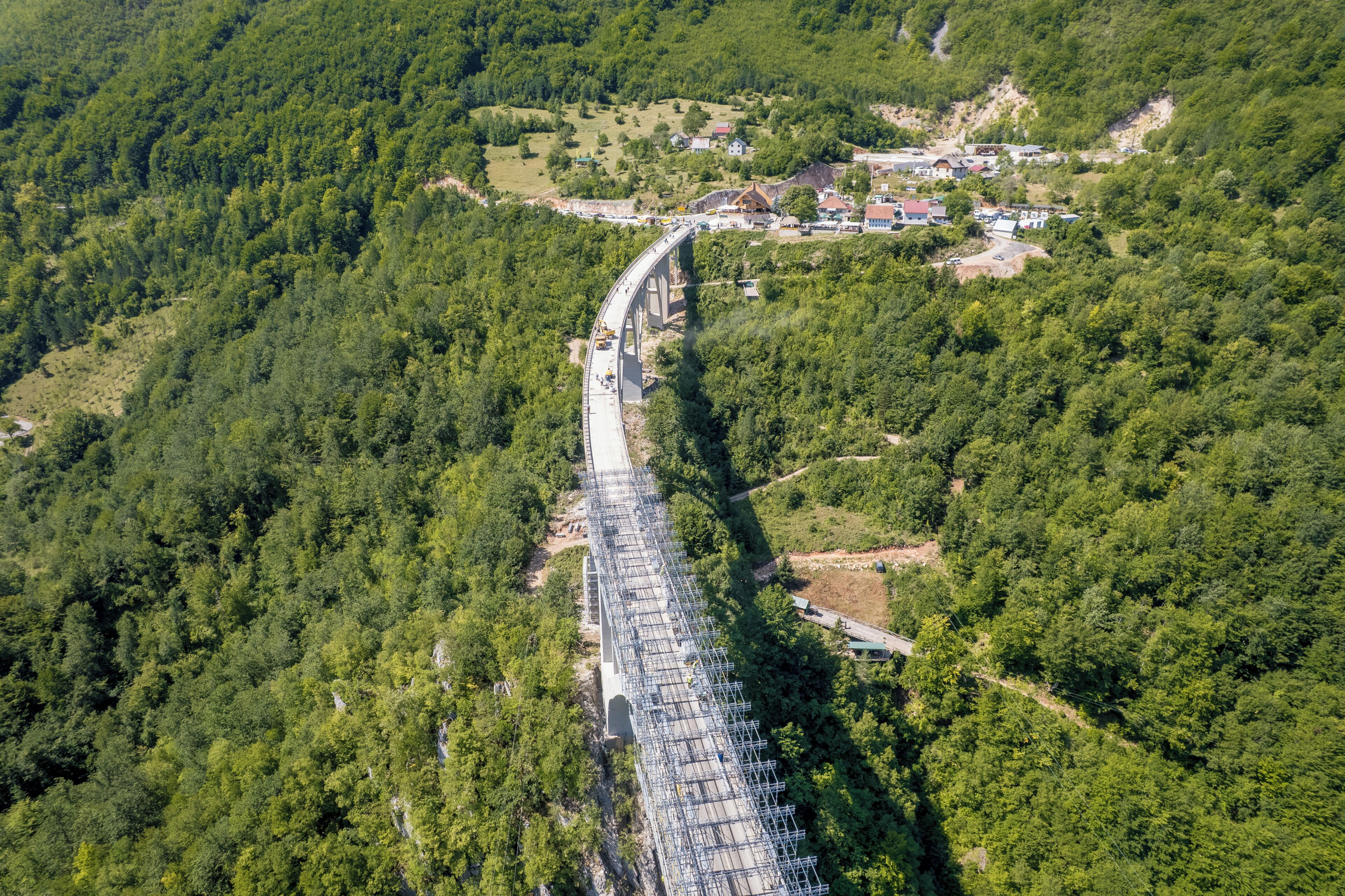 An aerial view of a bridge spanning a forested valley with a small settlement in the background.