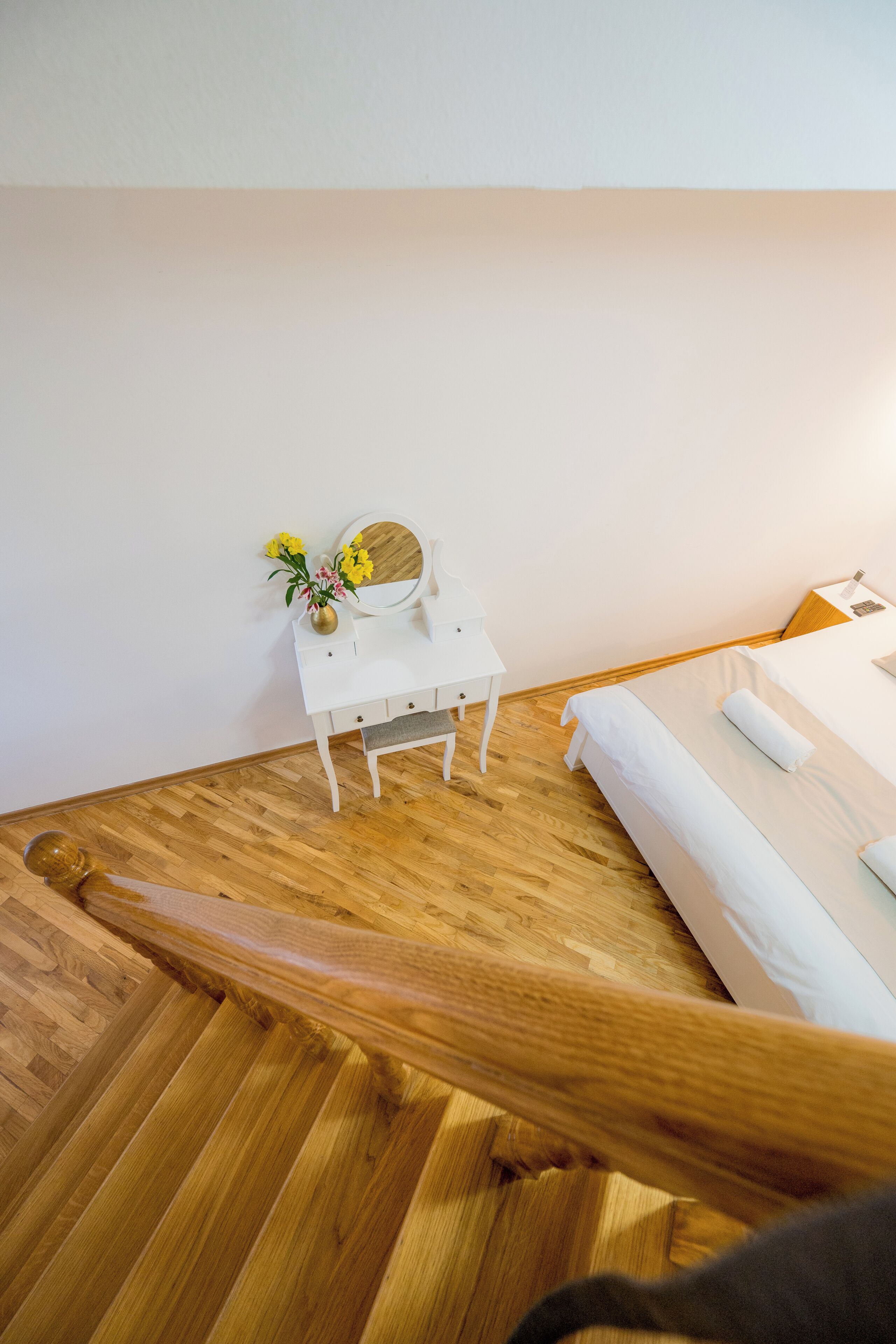 Ein Schlafzimmer mit Holzboden, einem Bett mit weißer Bettwäsche, einem kleinen weißen Nachttisch mit Blumen in einer Vase und einem runden Spiegel.