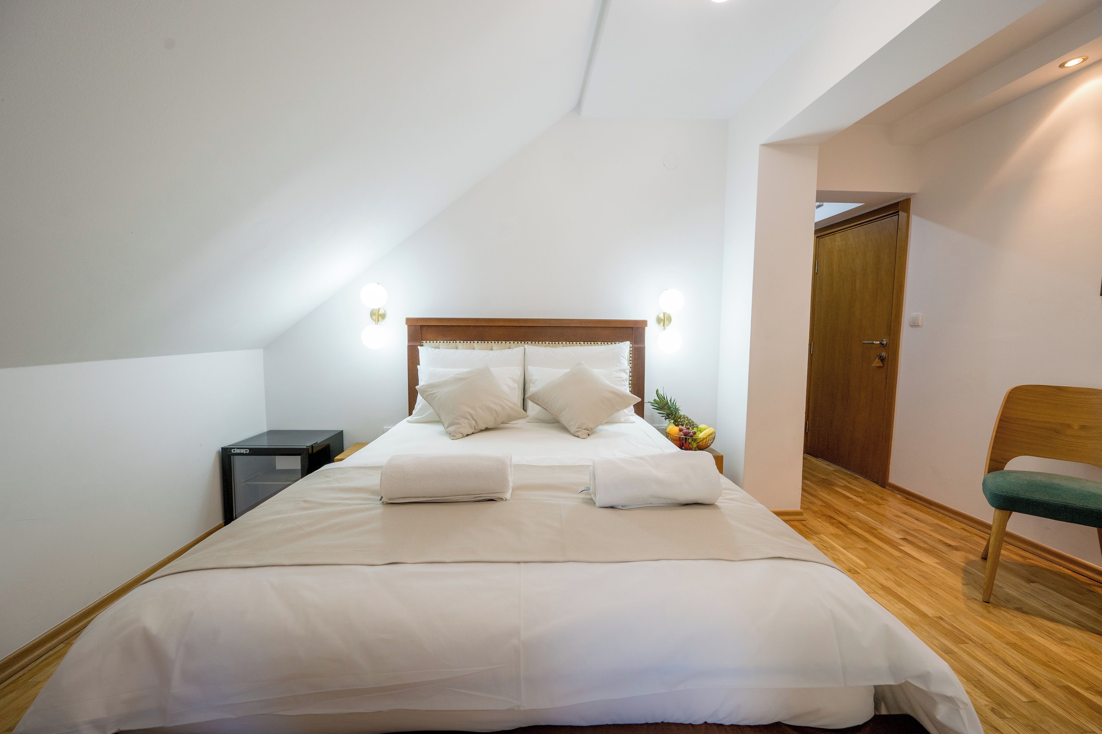 Una cama bien hecha con almohadas y una toalla doblada, una mesita de noche con una lámpara, y un suelo de madera.