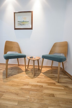 Two wooden chairs with blue cushions and a small wooden table with a remote control, against a white wall with a framed picture.
