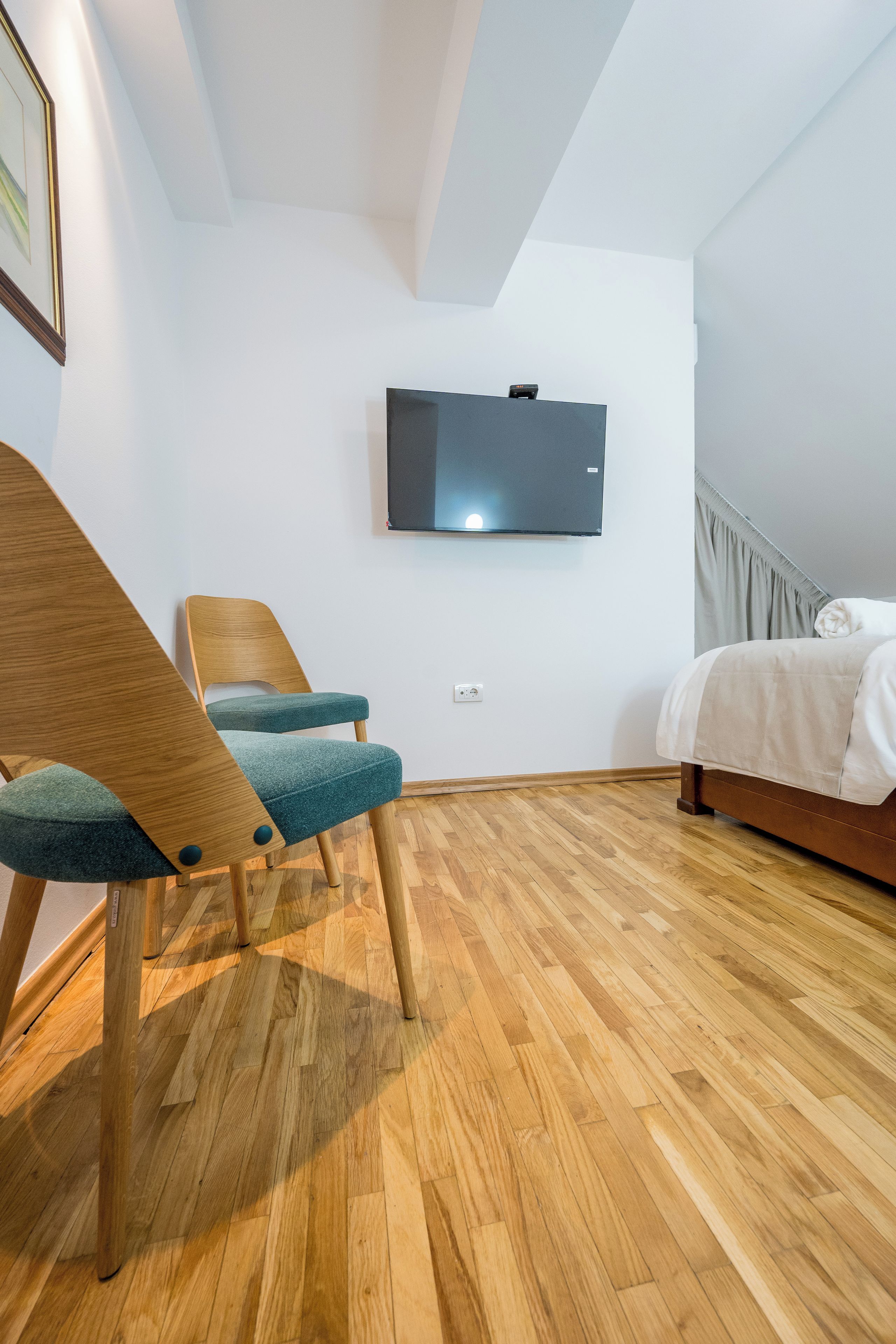 A modern bedroom with a wooden floor, a bed, a chair, and a flat-screen TV mounted on the wall.