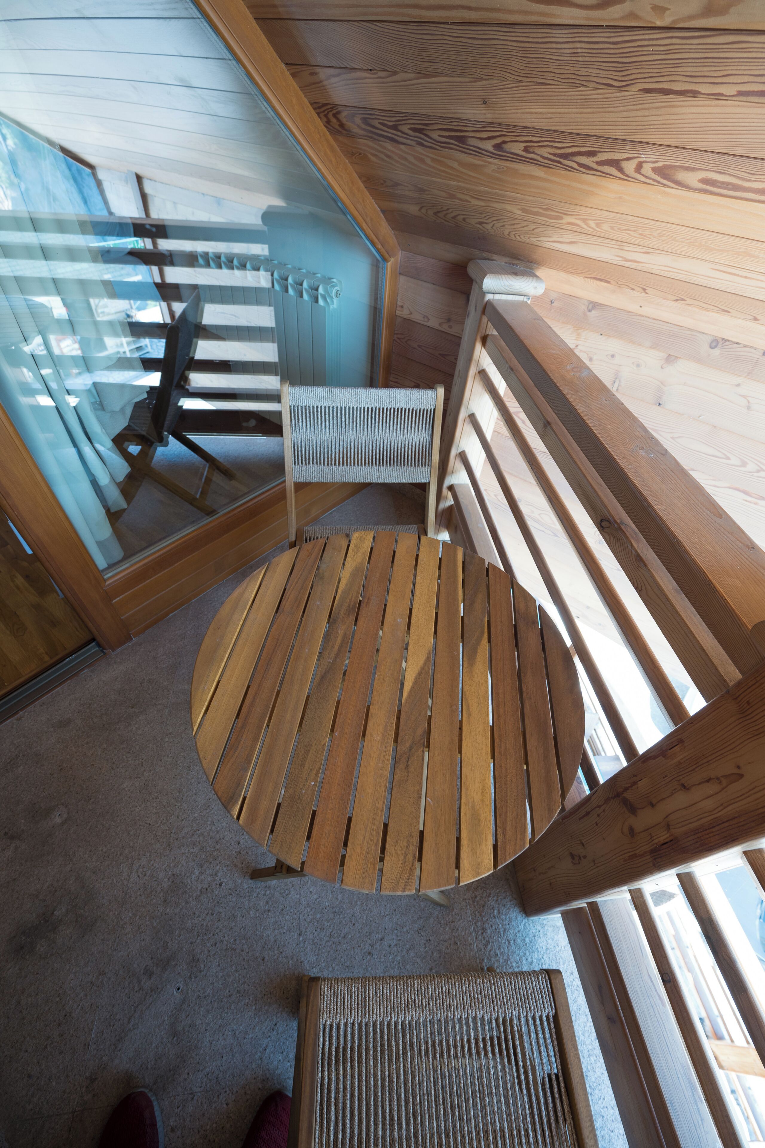 Una terraza de madera con una mesa redonda y una silla, un vidrio y vista al interior.