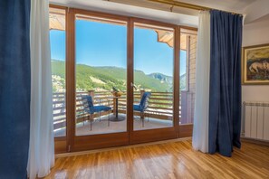 A room with a wooden floor, a sliding glass door leading to an outdoor balcony with two blue chairs and a table, and a painting on the wall.
