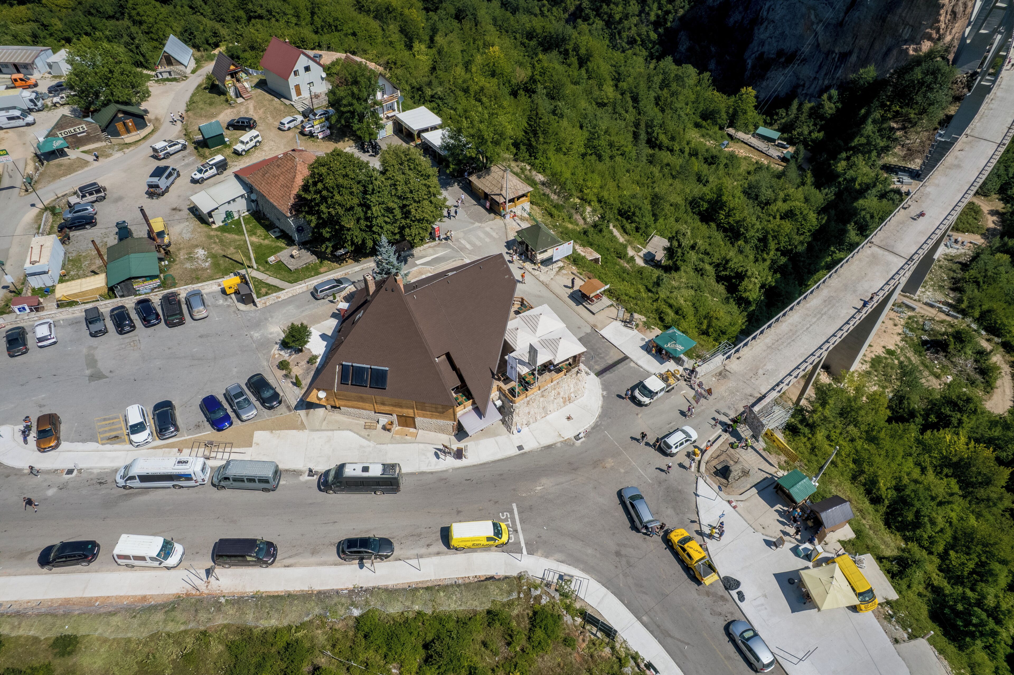 Une vue aérienne d’un parking où sont stationnés plusieurs véhicules, d’un bâtiment avec un toit rouge et d’un pont en arrière-plan.