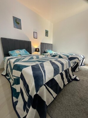Une chambre d’hôtel avec deux lits, une tête de lit grise, une petite table avec une lampe et un sol recouvert de moquette.