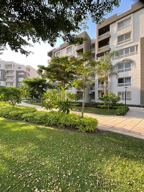 A multi-story residential building with balconies, surrounded by well-maintained greenery and a paved walkway. - Terrific 3-bedroom apartment with AC and Free-Wifi in lovely Madinty Garden View (Madinaty)
