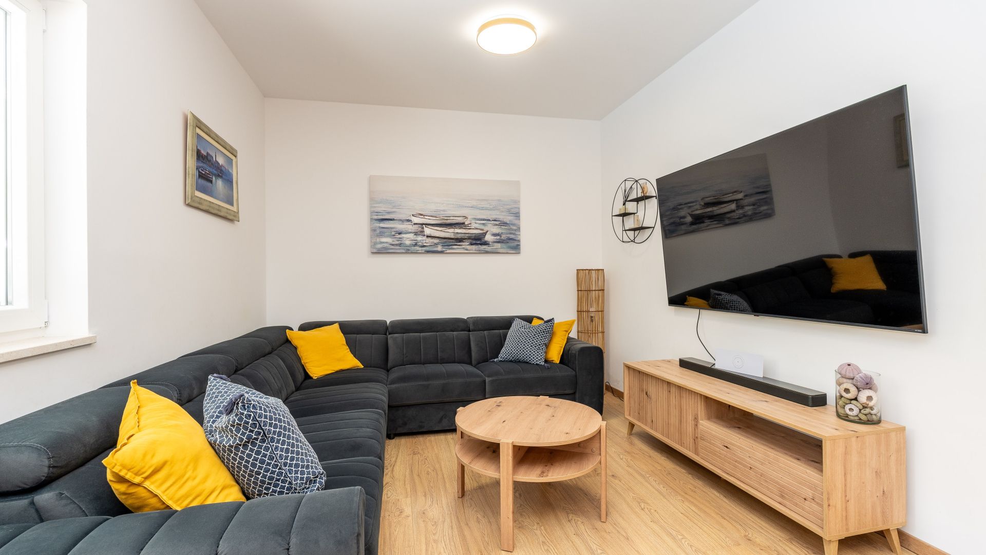 A modern living room with a sectional sofa, a round wooden table, a TV mounted on the wall, and a painting above the sofa.