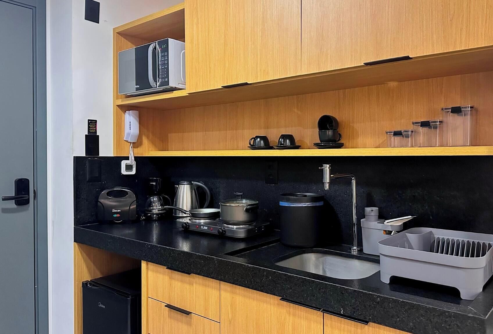 A compact kitchen with a black countertop, wooden cabinets, a coffee machine, a kettle, a coffee pot, a kettle, a coffee maker, a sink, and a dish rack.