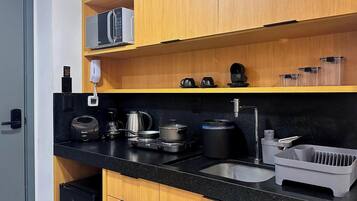 A compact kitchen with a black countertop, wooden cabinets, a coffee machine, a kettle, a coffee pot, a kettle, a coffee maker, a sink, and a dish rack.