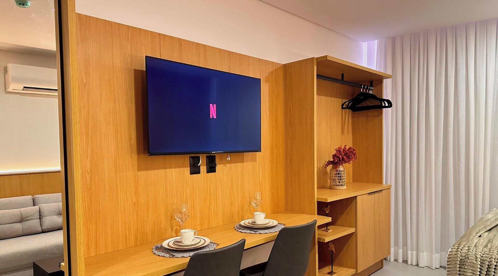 A modern living room with a flat-screen TV mounted on a wooden wall, a dining area with a table set for two, and a built-in shelving unit.