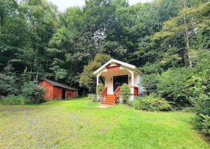 A small wooden structure with a porch and steps, surrounded by lush greenery and a red barn in the background. - Creekfront • Pet Friendly • Sleeps 4 • Near Boone & West Jefferson (Todd)