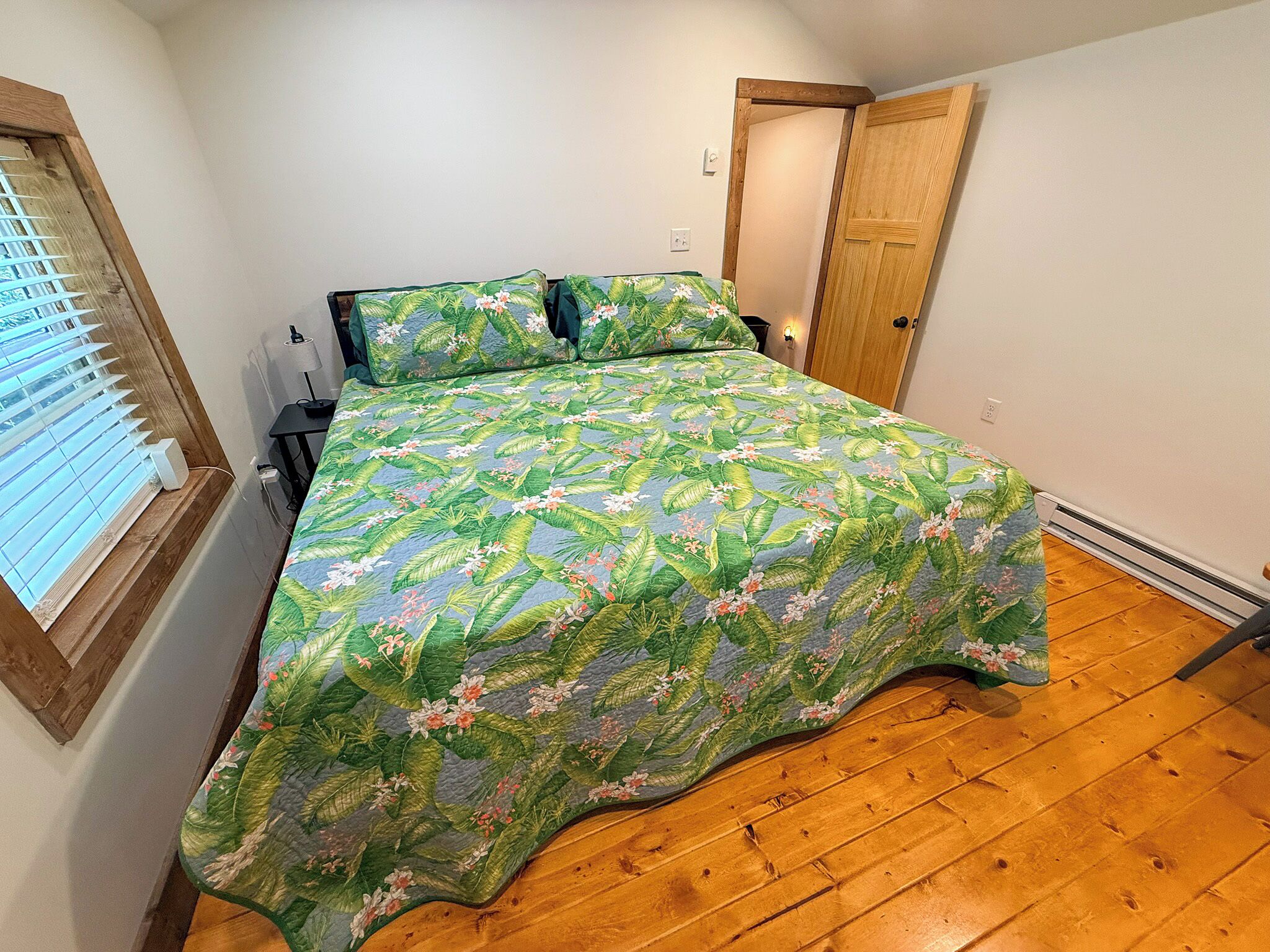A bedroom with a bed, a window with blinds, a door, and a wooden floor.
