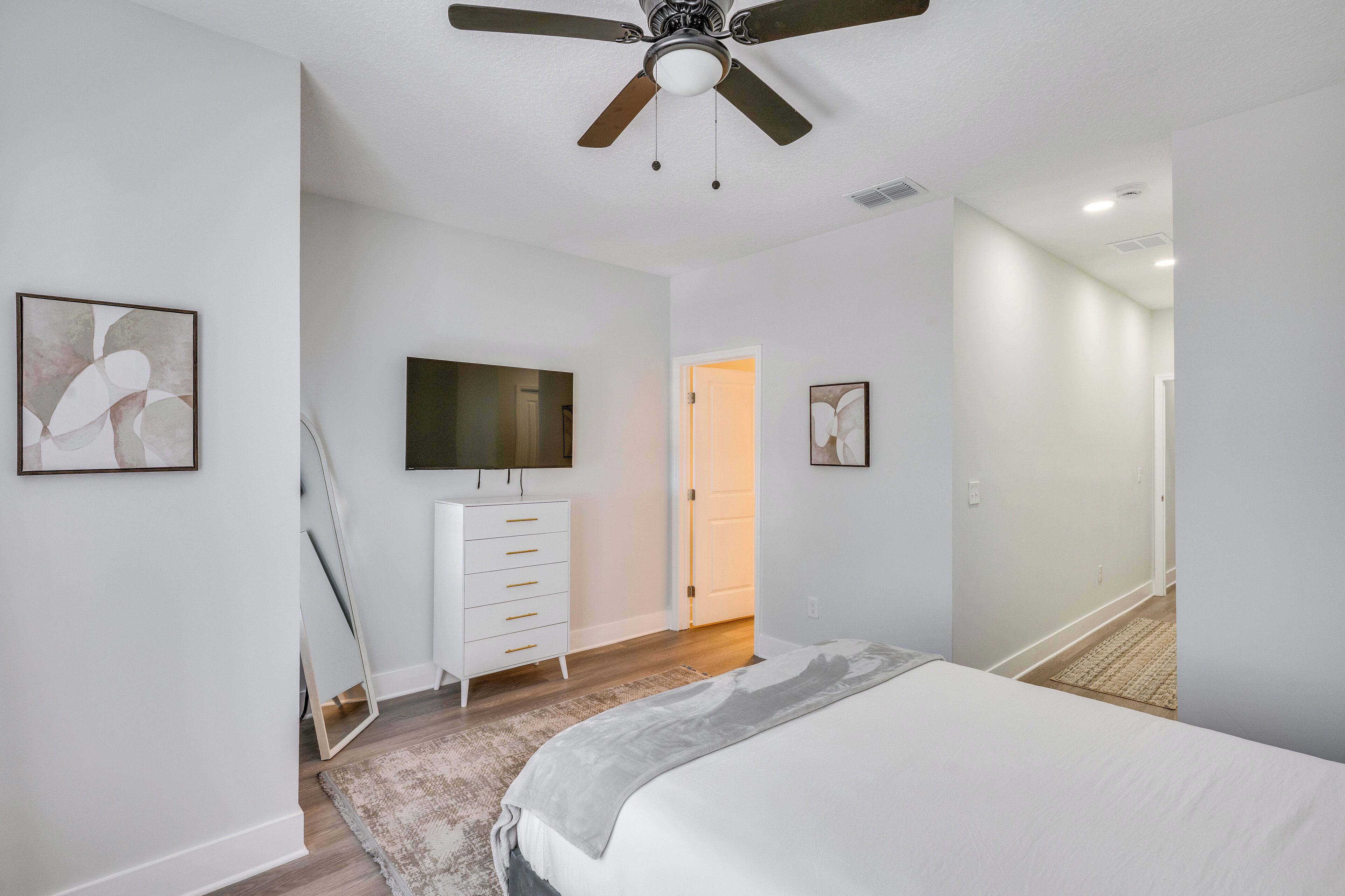 A bedroom with a bed, a dresser, a ceiling fan, and artwork on the walls.