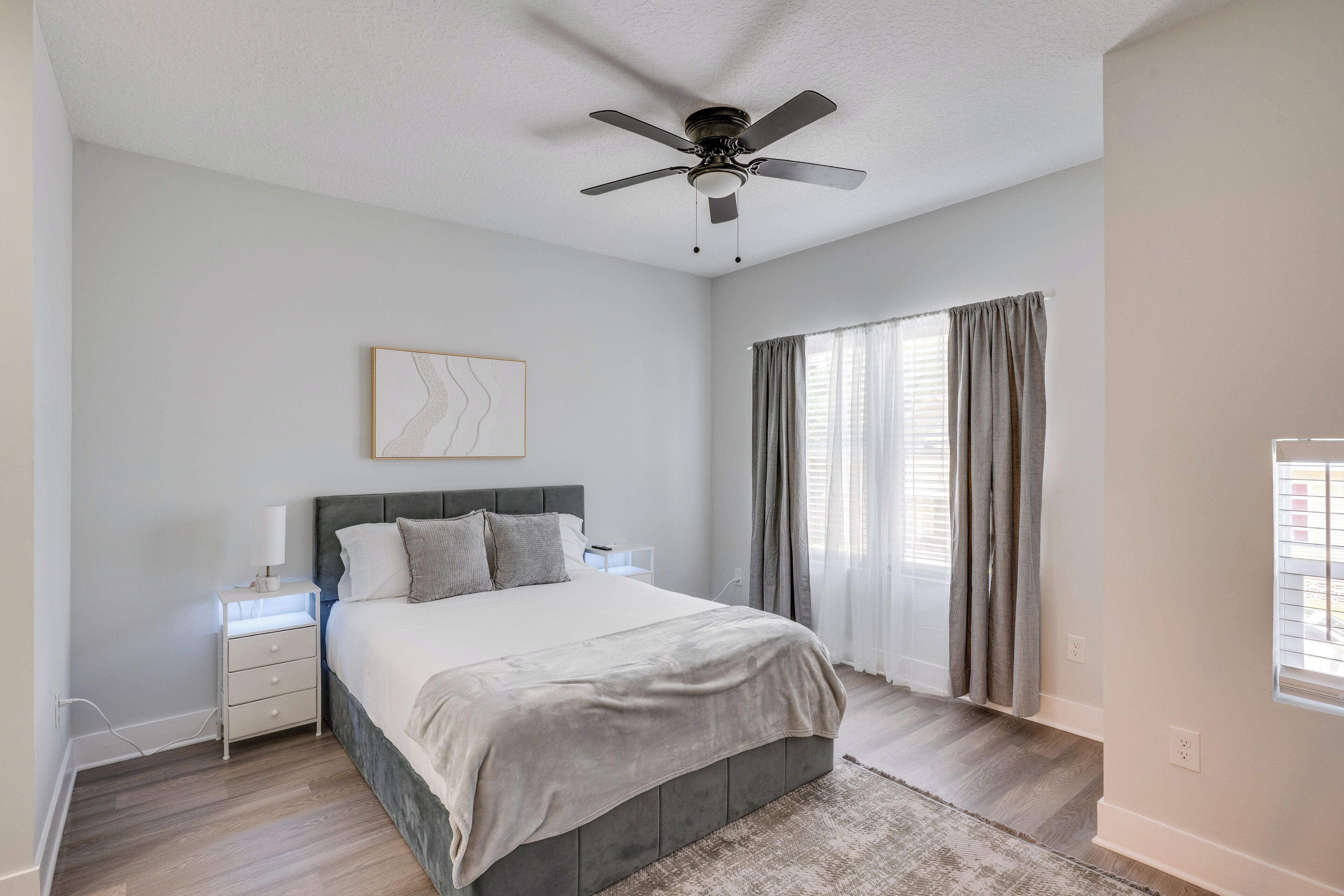 A bedroom with a bed, nightstands, a ceiling fan, a window with curtains, and a wall-mounted artwork.