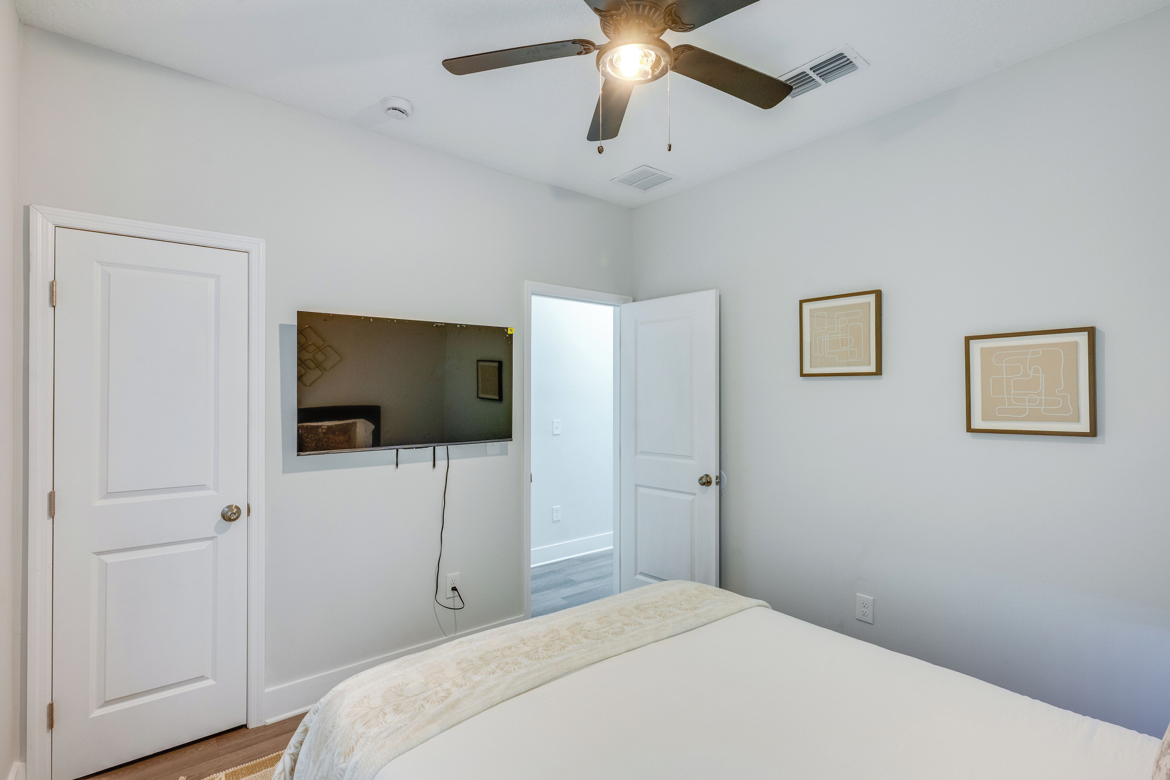 A bedroom with a ceiling fan, a bed with a white bedspread, two doors, and two framed pictures on the wall.