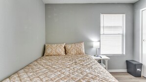 A bedroom with a bed, a nightstand, a lamp, a window, and a trash bin. - 2-unit Combo 10 Mi to Vintage Park & Airport! (Houston)
