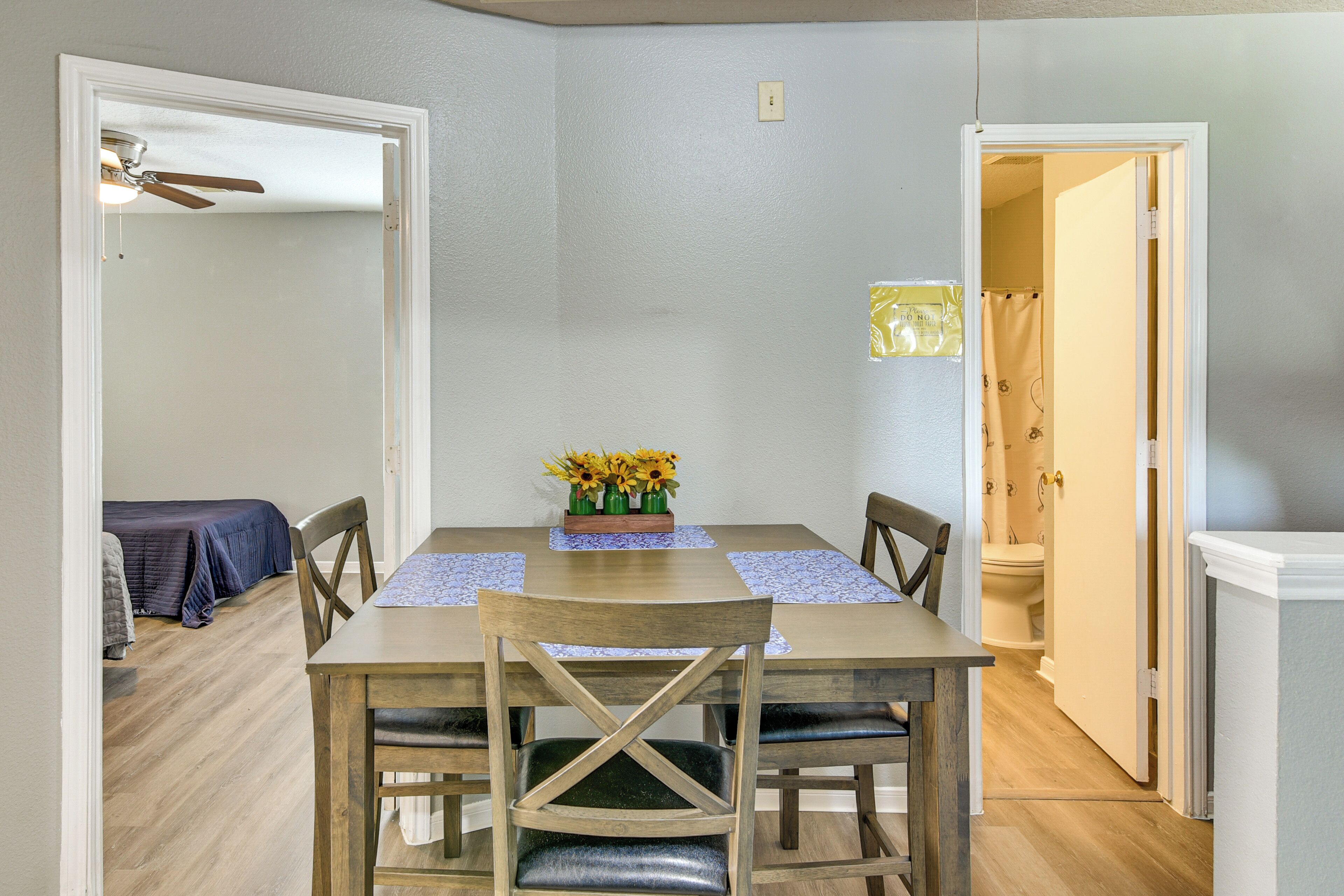 Un coin repas avec une table, des chaises et un vase de fleurs. Un ventilateur de plafond est installé au-dessus de la table, et une porte donne accès à une autre pièce.