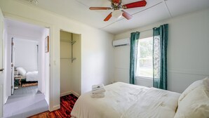 A bedroom with a bed, a ceiling fan, an air conditioning unit, a window with curtains, and a door leading to another room. - Pet-friendly Cottage Near Beach: King Bed/fire Pit (Bonita Springs)