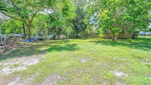 A backyard with a grassy lawn, trees, a wooden fence, and a pile of bricks. - Pet-friendly Cottage Near Beach: King Bed/fire Pit (Bonita Springs)