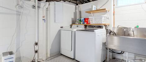 A laundry room with two washing machines, a sink, and a shelf with laundry supplies.