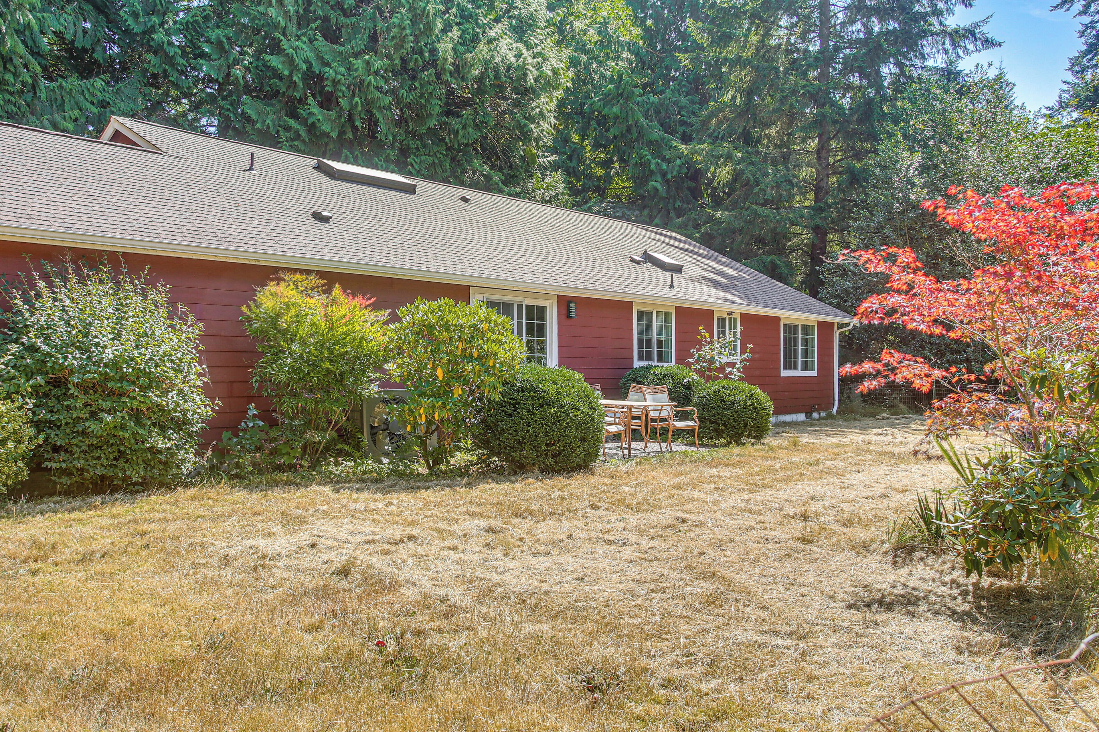 A red house with a sloped roof, surrounded by a dry grassy yard and various shrubs.