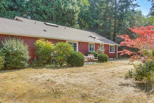 A red house with a sloped roof, surrounded by a dry grassy yard and various shrubs.