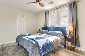 Un dormitorio con cama, mesita de noche, lámpara, ventilador de techo y ventana con cortinas.