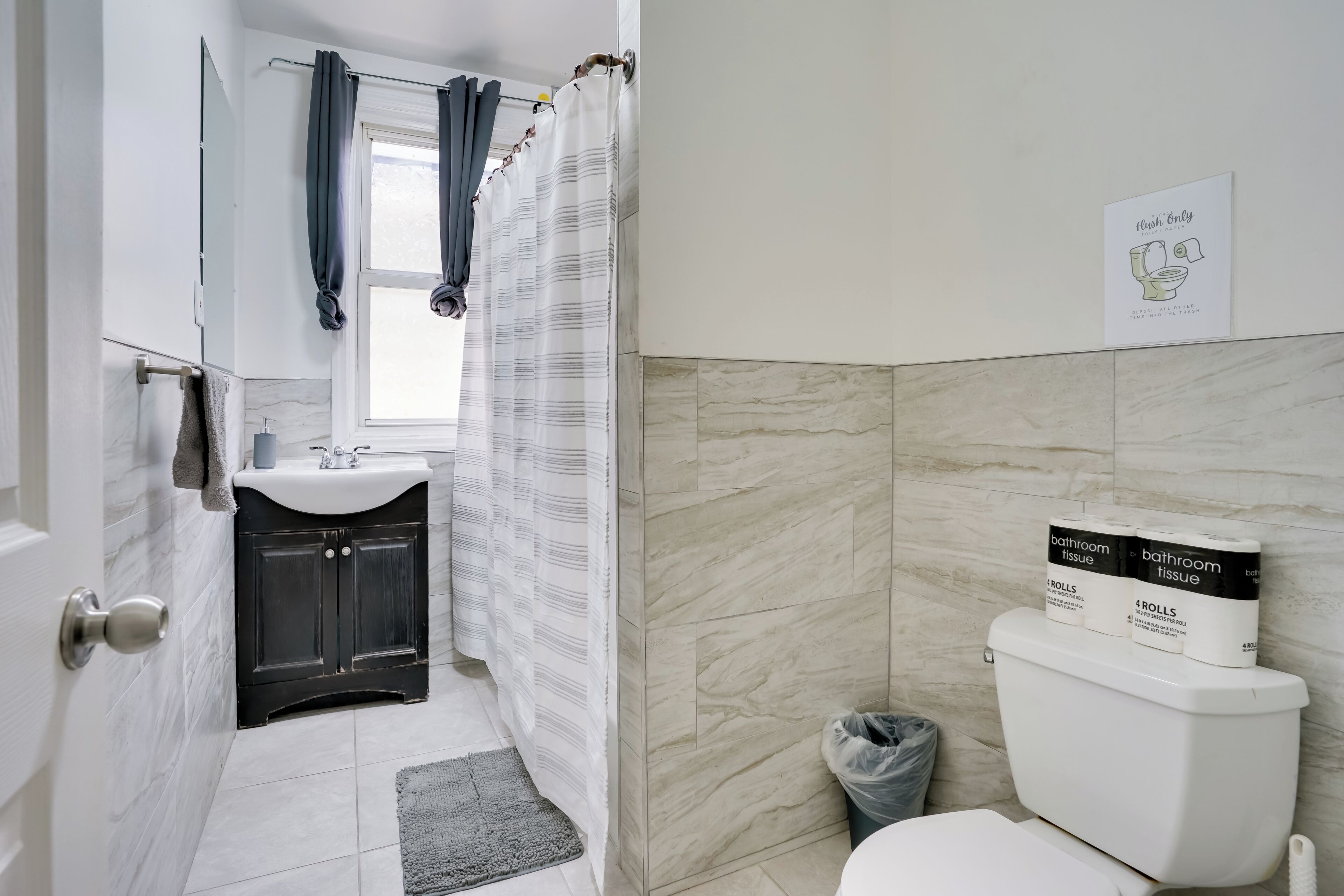 A bathroom with a toilet, a sink, a mirror, and a shower curtain. There are rolls of toilet paper and a trash bin.