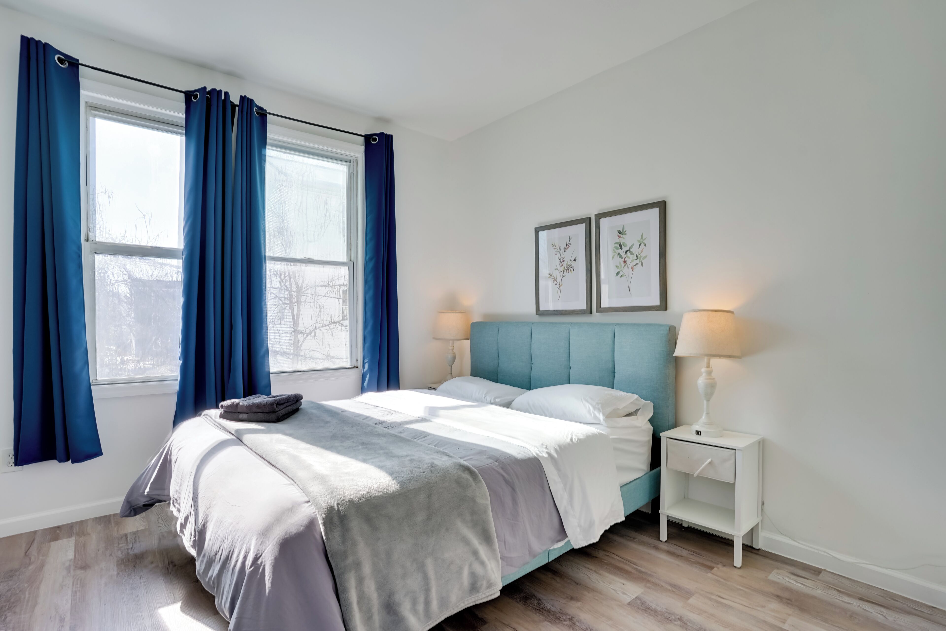 A bedroom with a bed, two bedside tables, a window with curtains, and wall art.