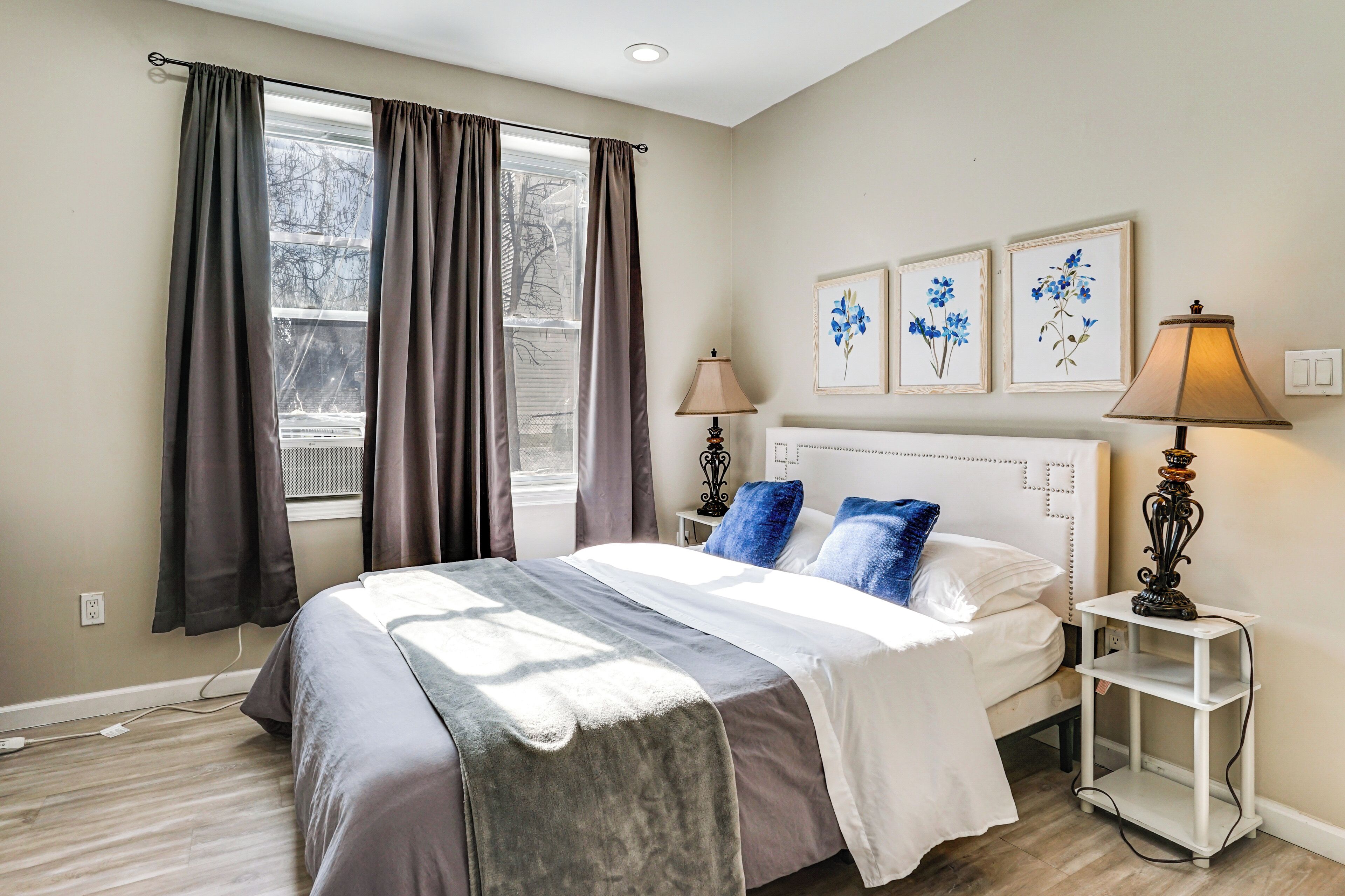 Een slaapkamer met een bed, twee bedlampen, een nachtkastje, een raam met gordijnen, en twee bloemschilderijen aan de muur.