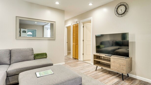 A modern living room with a grey sofa, a flat-screen TV on a wooden stand, and a mirror on the wall. - Updated Duplex - 3 Mi to Newark Penn Station! (Newark)