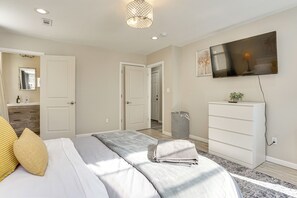 Quarto com cama, televisão na parede, cômoda e lixeira.