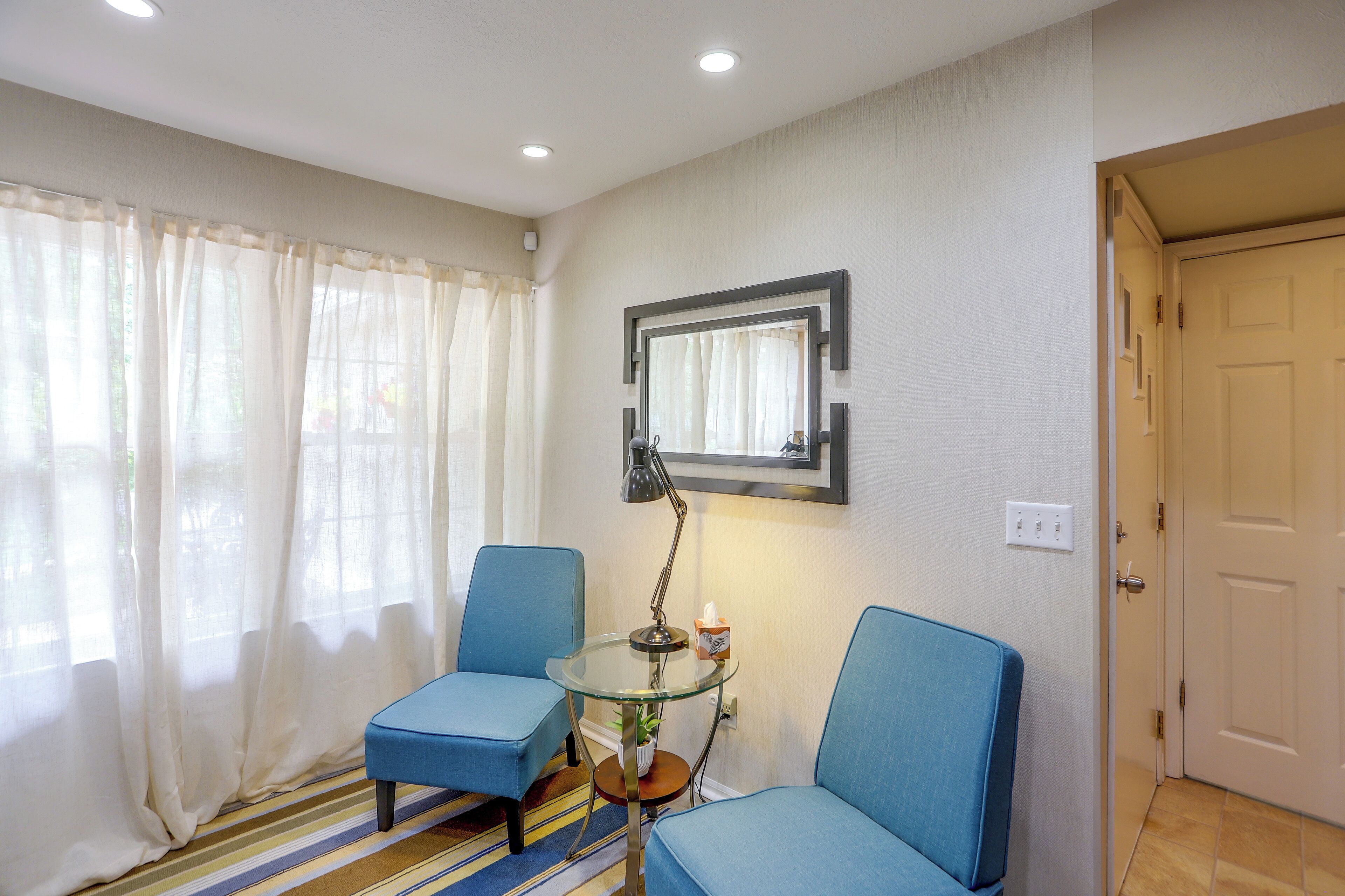 A room with two blue chairs, a small glass table with a lamp, a mirror on the wall, and a door.