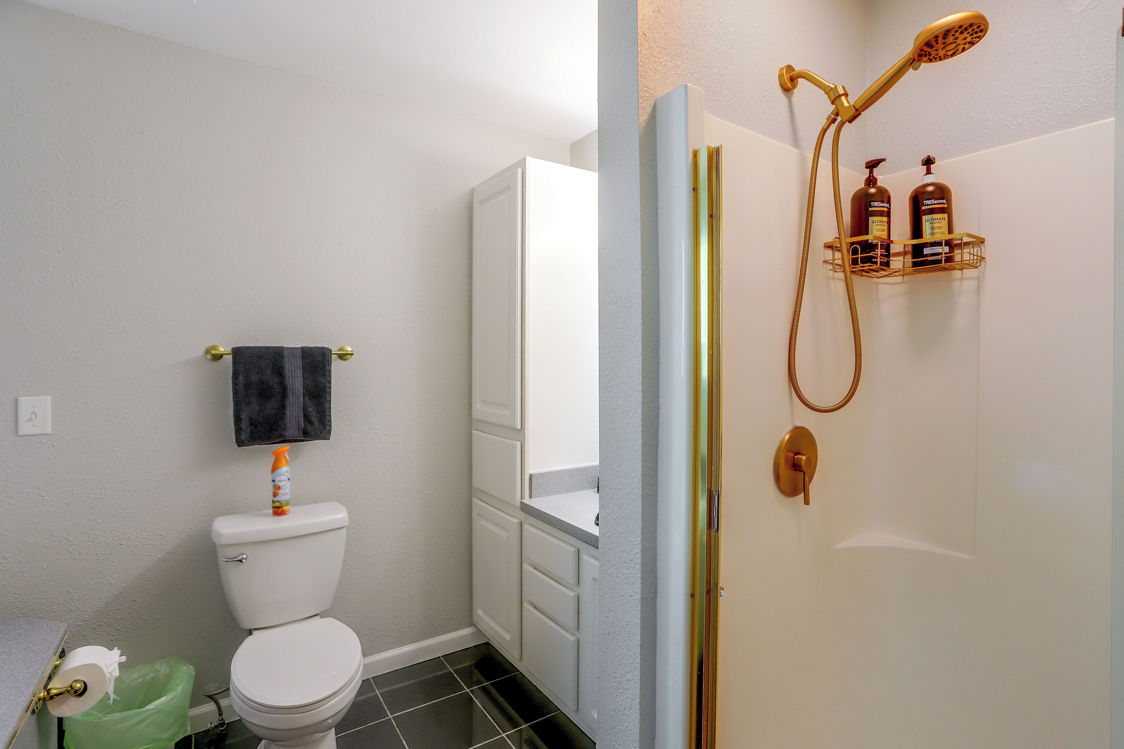 A bathroom with a toilet, a towel rack, a shower with a gold showerhead, and toiletries on a shelf.