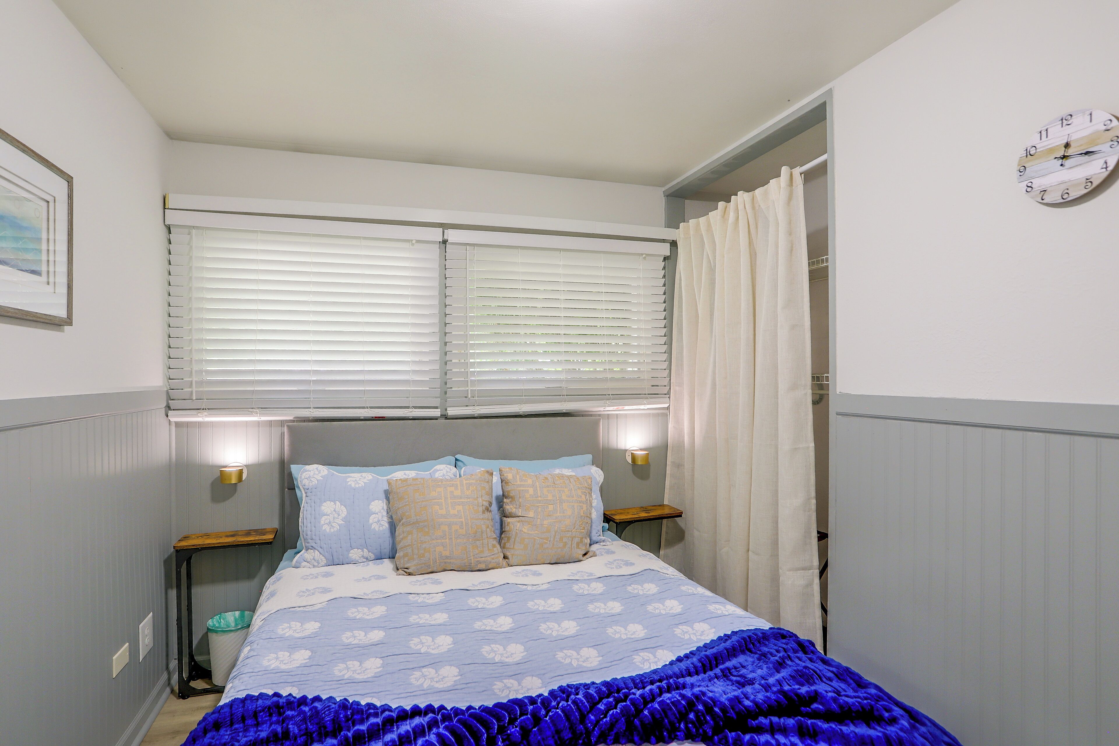 A bedroom with a bed, two bedside tables, a wall clock, and a window with blinds.