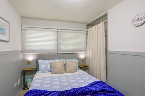 A bedroom with a bed, two bedside tables, a wall clock, and a window with blinds.