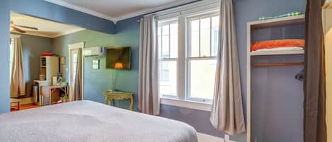 A bedroom with a bed, a ceiling fan, a window with curtains, a shelf with towels, and a desk with a lamp.