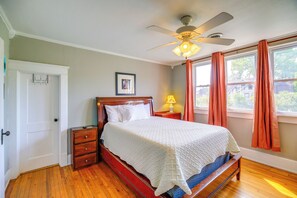 Un dormitorio con una cama de madera, un ventilador de techo, una mesita de noche con lámpara, una puerta y una ventana con cortinas.