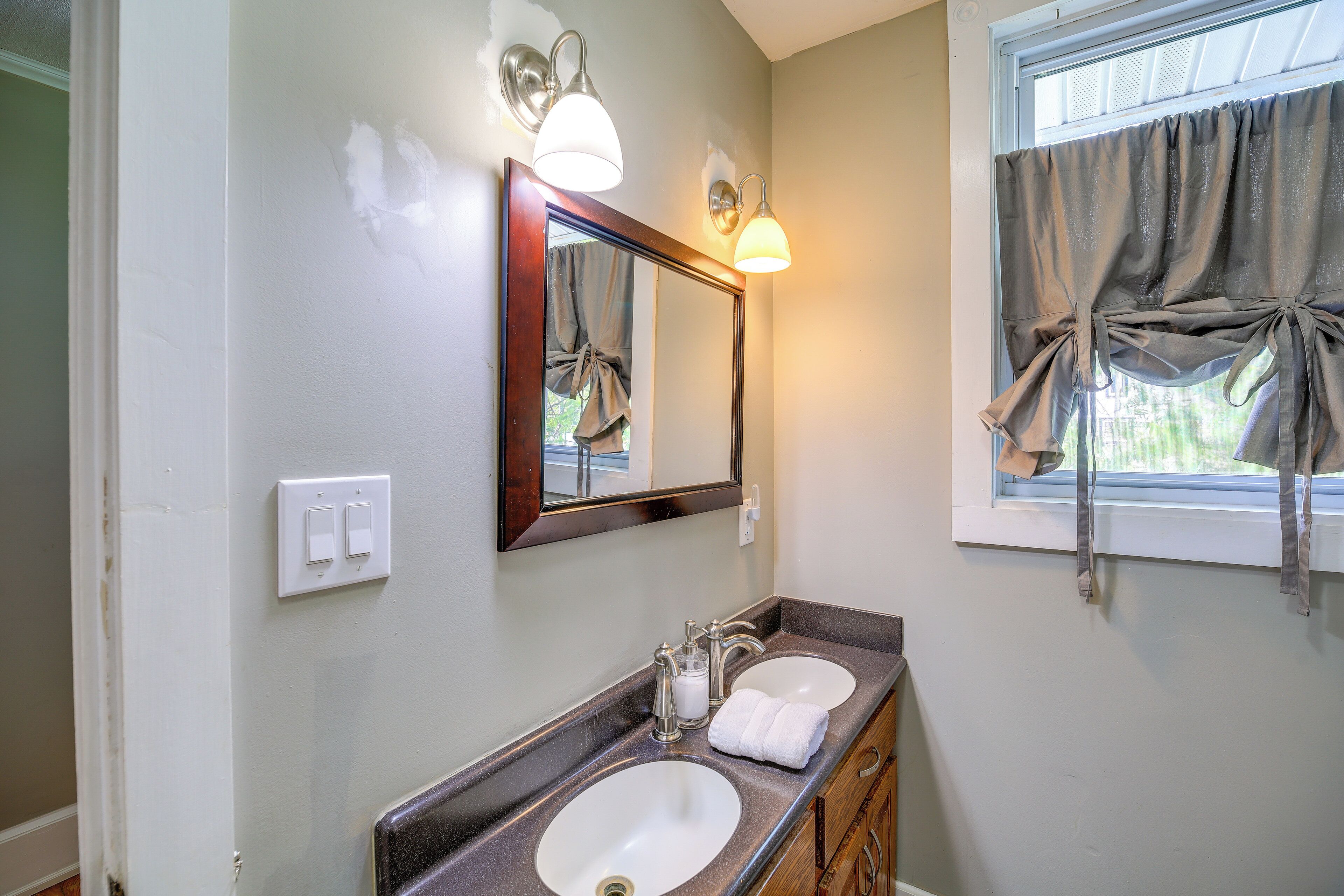 A bathroom with a double sink, a mirror, wall-mounted lights, and a window with curtains.