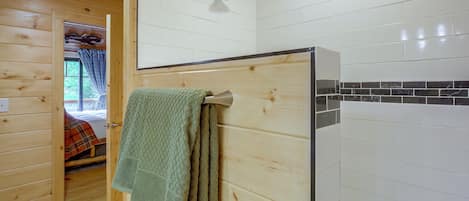 A wooden interior with a shower, a towel hanging on the wall, and a glimpse of a bed with a plaid blanket.