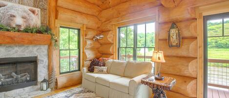 A log cabin interior with a fireplace, a sofa, a lamp, and a view of the outdoors.