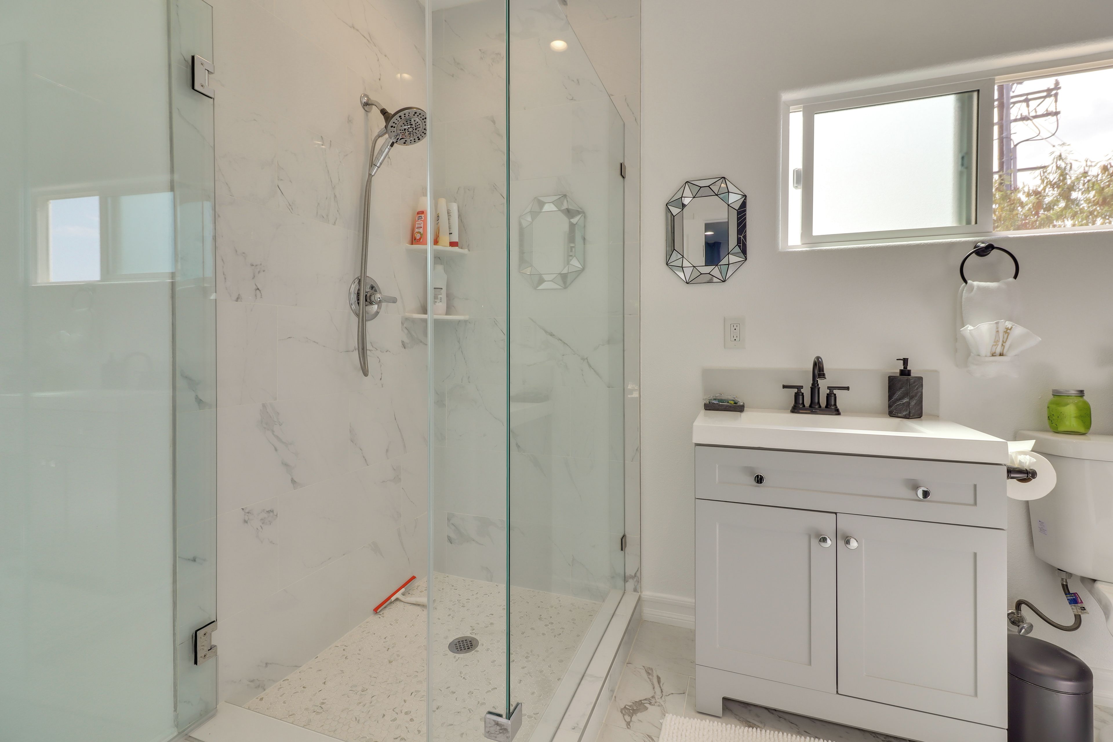 Un baño moderno con ducha envuelta en vidrio, un tocador blanco con espejo y una ventana sobre el lavamanos.