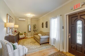 A living room with a sofa, a chair, a lamp, a small table, and an air conditioning unit. There is a door with an exit sign above it.