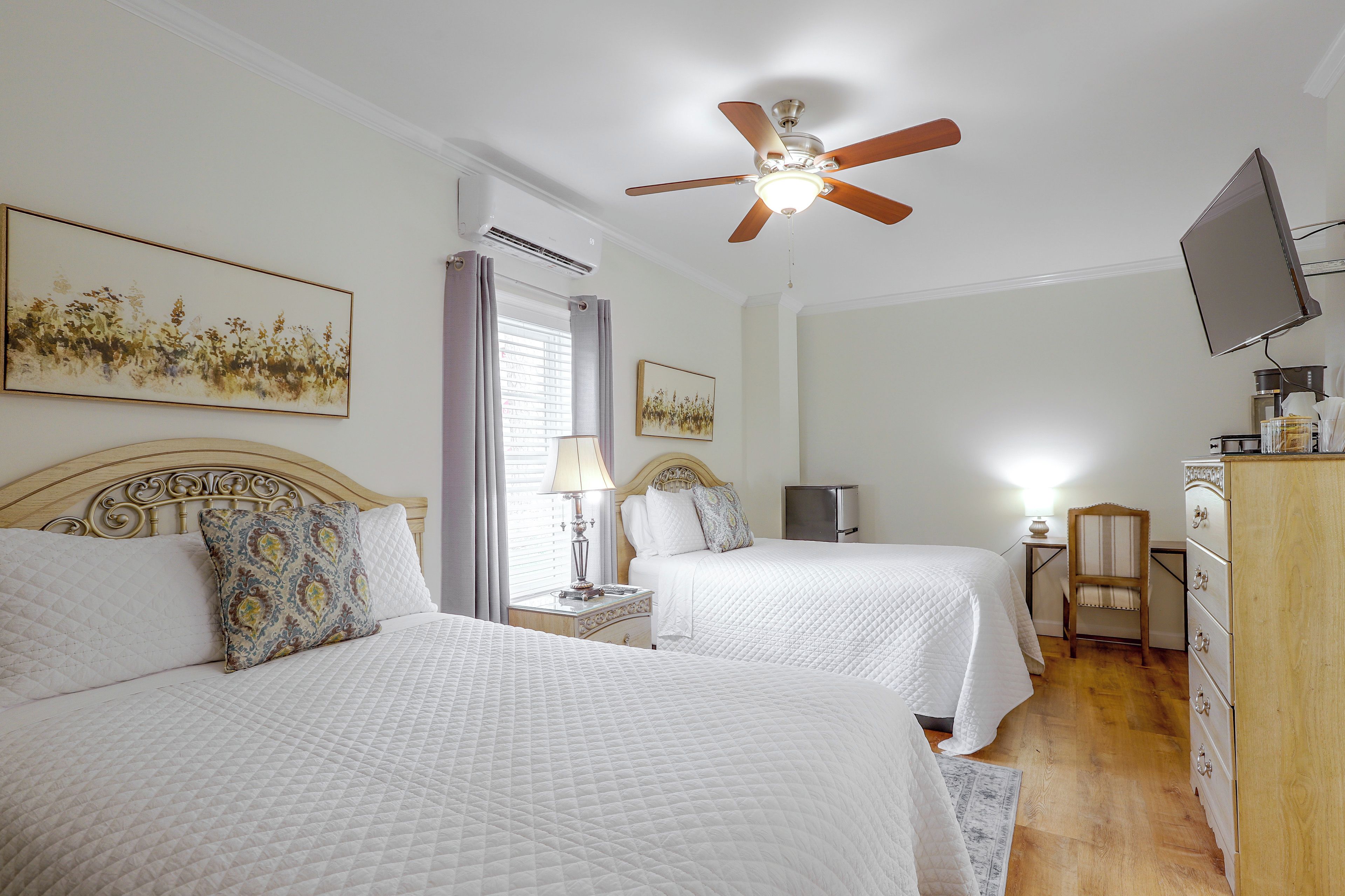 A hotel room with two beds, a ceiling fan, a television, and artwork on the walls.