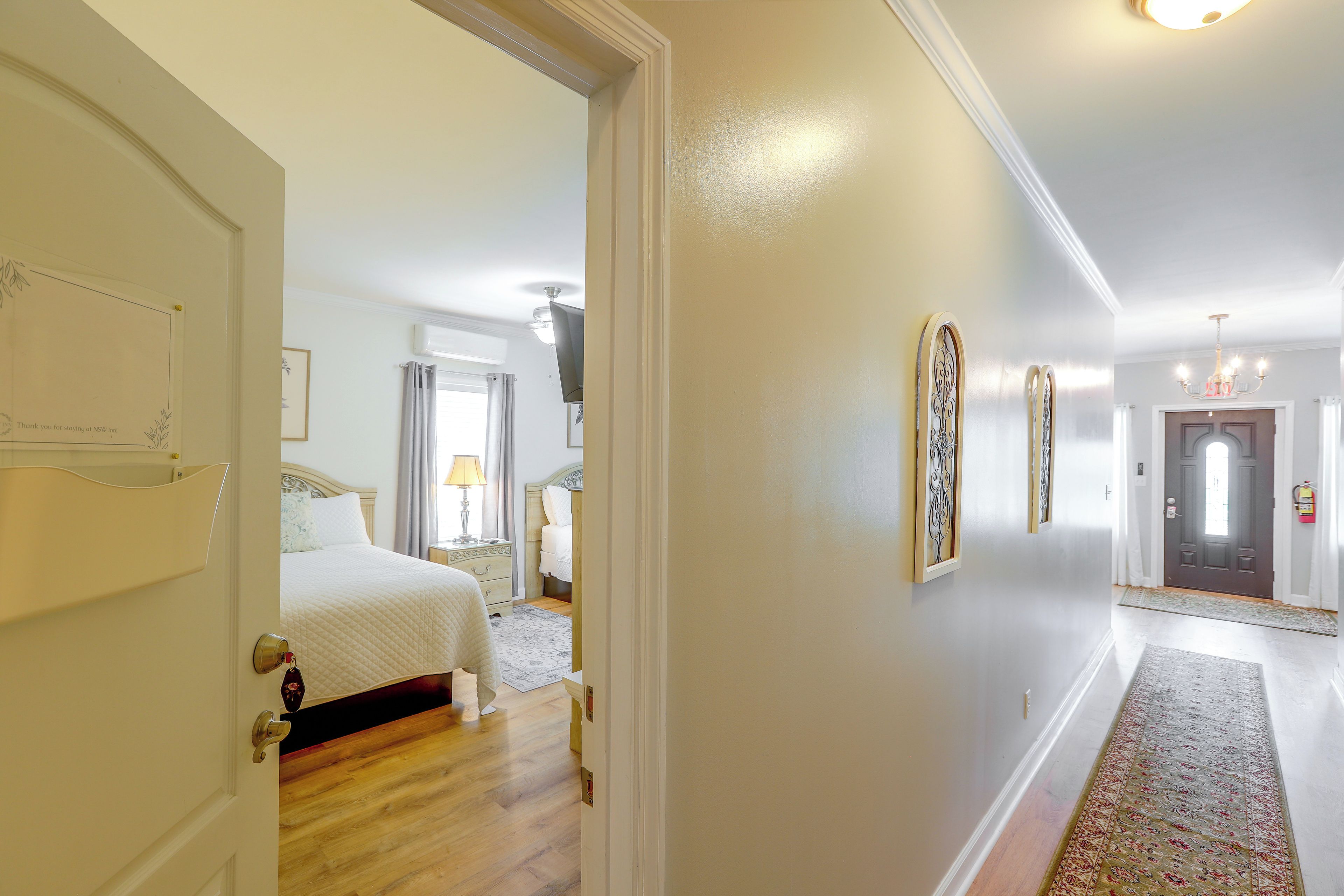 Un couloir d’hôtel avec un lit, une table de chevet, une lampe et une porte donnant sur une autre pièce.