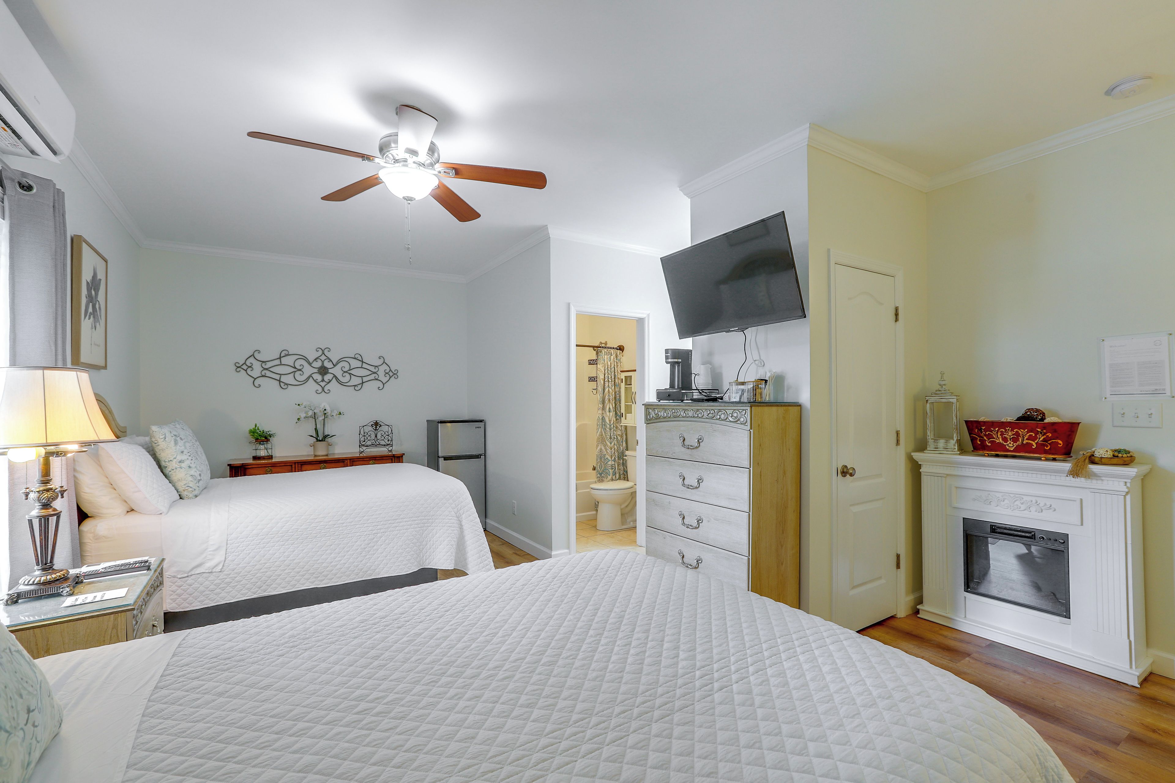 A bedroom with two beds, a ceiling fan, a television, a fireplace, and a door leading to another room.