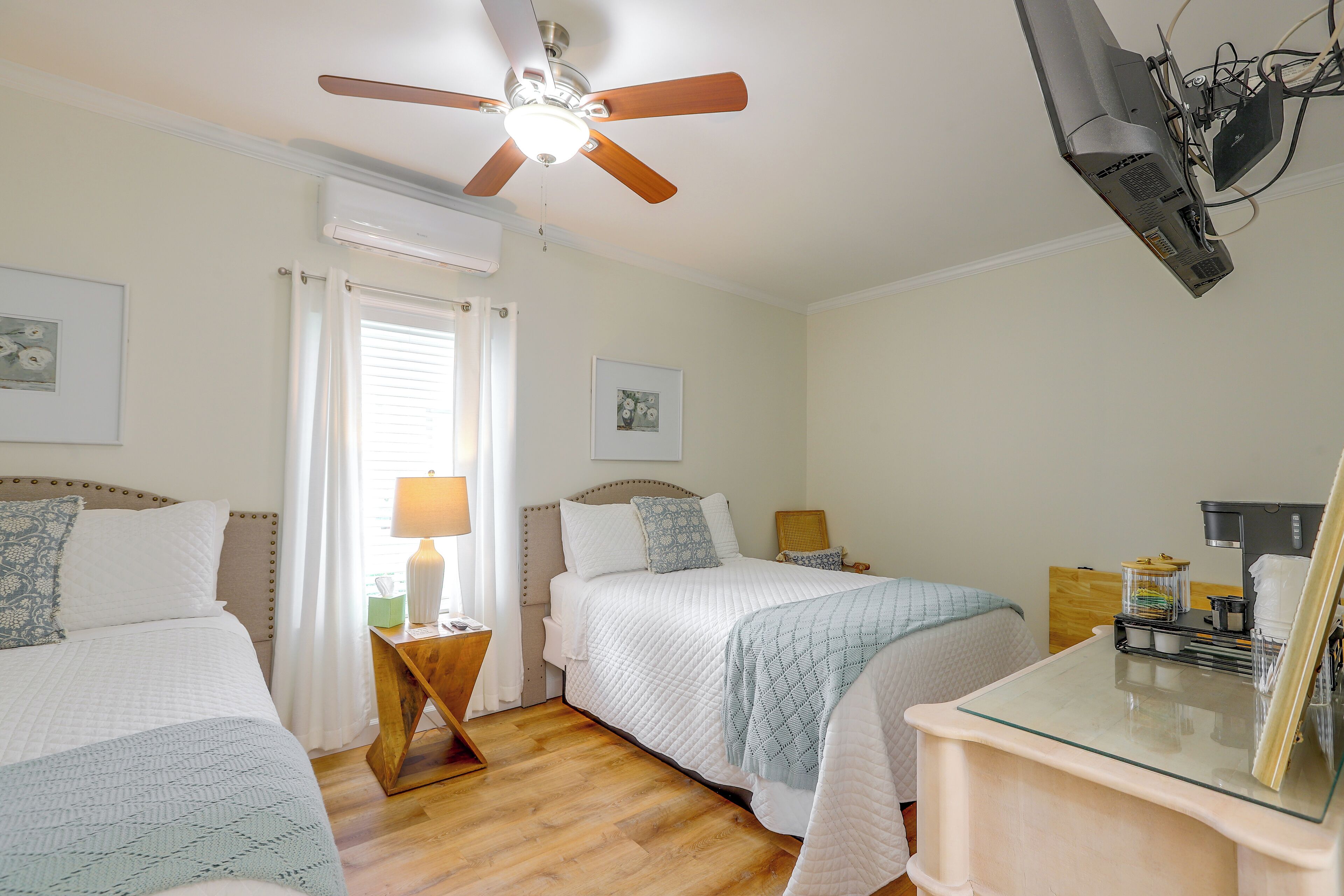 A hotel room with two beds, a ceiling fan, a desk with a coffee maker, and a window with curtains.
